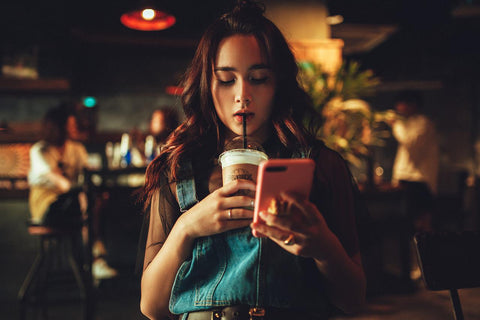 A women checking her phone while drinking a drink like one you may find in a Starbucks. She appears to be in a coffee shop. 