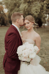 Groom in a sharp burgundy suit holds a stunning bouquet of white roses, featuring the exquisite floral arrangements of Floret and Foliage - Fargo's premier wedding florist