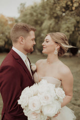 Groom in a sharp burgundy suit holds a stunning bouquet of white roses, featuring the exquisite floral arrangements of Floret and Foliage - Fargo's premier wedding florist