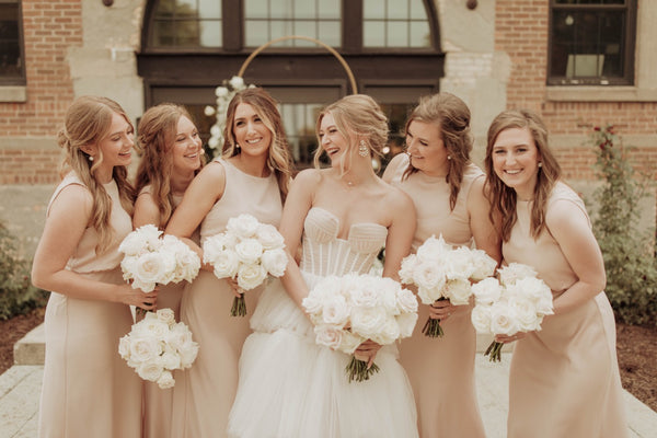 Bridal party with bouquets dresses in blush bridesmaids dresses and holding white and blush rose bouquets in front of a ceremony arch 