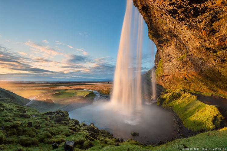 An image from behind and off to the side of a very big waterfall. The water falls from an overhanging cliff face where it gathers in a pool surrounded by short dense grass before flowing off in a river through a flat landscape.