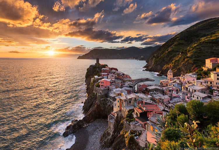 An image from a hill top looking down on a quaint italian mediterranian seaside village. The cloudy sky is lit up near sunset as the vast sea breaks against the shore