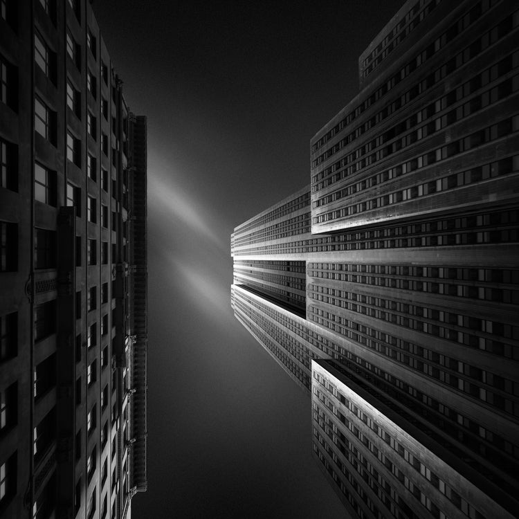 A colorless image looking straight up between two syscraper buildings at the sky, one lit up from the sun, the other in shadow, a faint wisp of a cloud can be seen