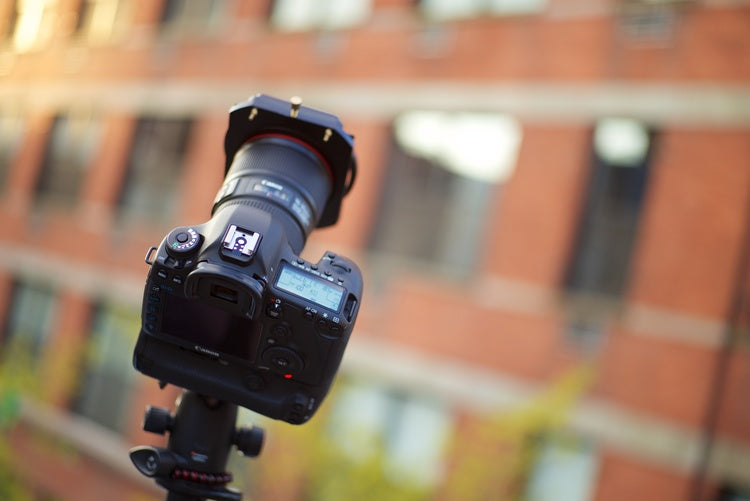 An image of a DSLR style camera and lens with a Formatt Hitech filter holder and filter attached to the front. It is mounted to a tripod and pointed at the sky with a brick building in the background
