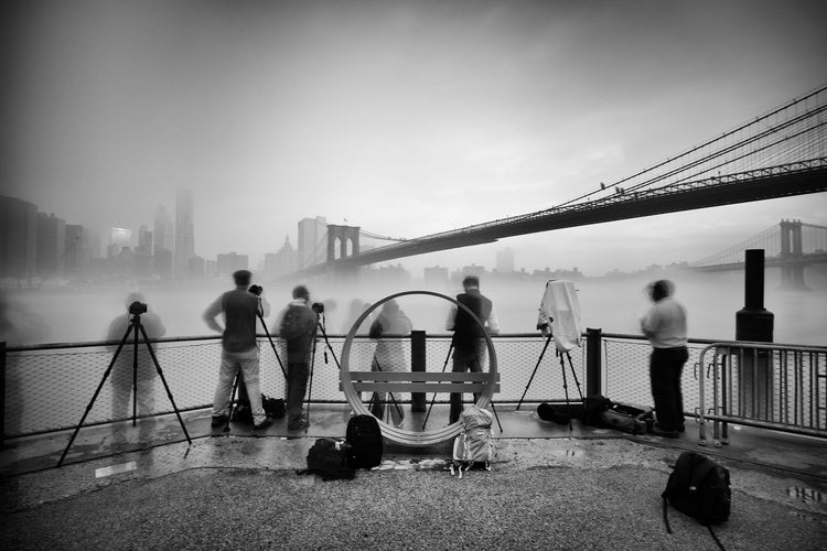 An old suspension bridge over a river with heavy fog separating the blurry image of people in the foreground