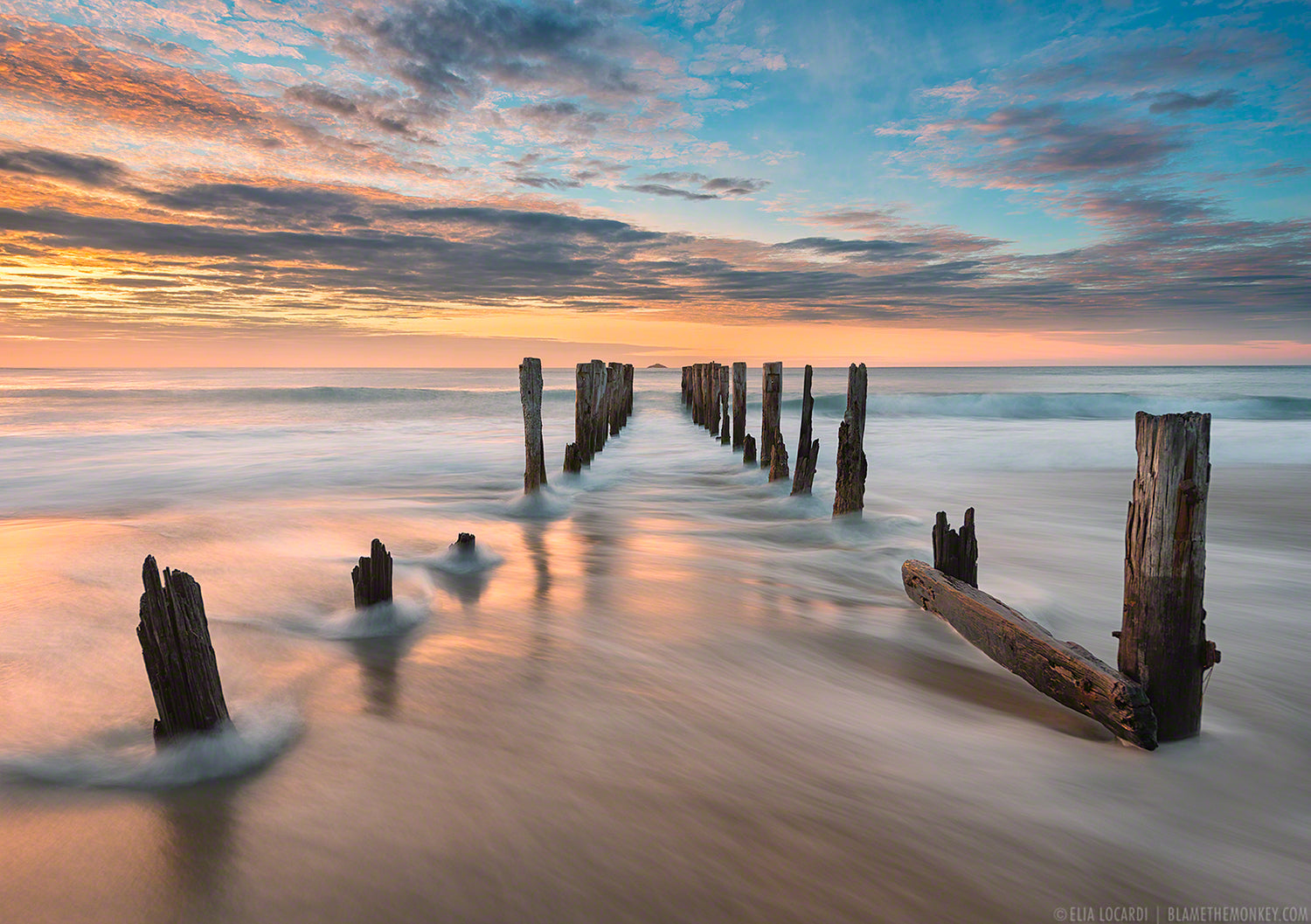 An ocean sunset of blurry waves in motion through the rotting wooden pylons of a collapsed dock
