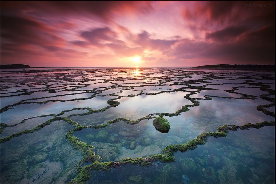 An image shot across a large area of low tide-pools framed by two slivers of land fading off into the ocean at sunset