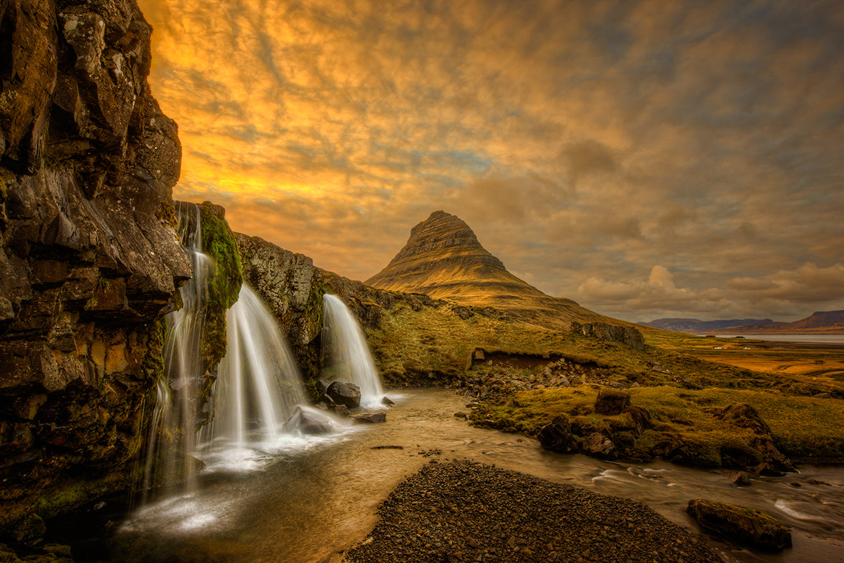 An image of Kirkjufellsfoss in Iceland with a very cloudy sky. There is a steep triangular grass covered mountain in the background with a waterfall in the foreground.