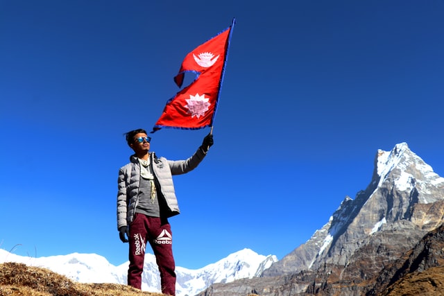 Man standing in a mountain scene holding the flag of Nepal