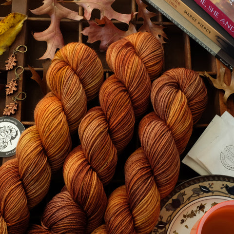 Autumnal brown and orange variegated yarn shown with oak leaf stitch markers, tea, a sticker, and books among leaves on a wooden tray.