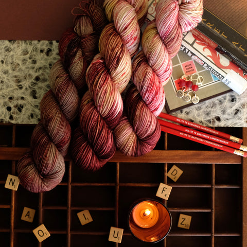Red variegated yarn with blush and peach hues inspired by Margaret Atwood's work, amongst scrabble tiles, Atwood books, and themed extra gifts.