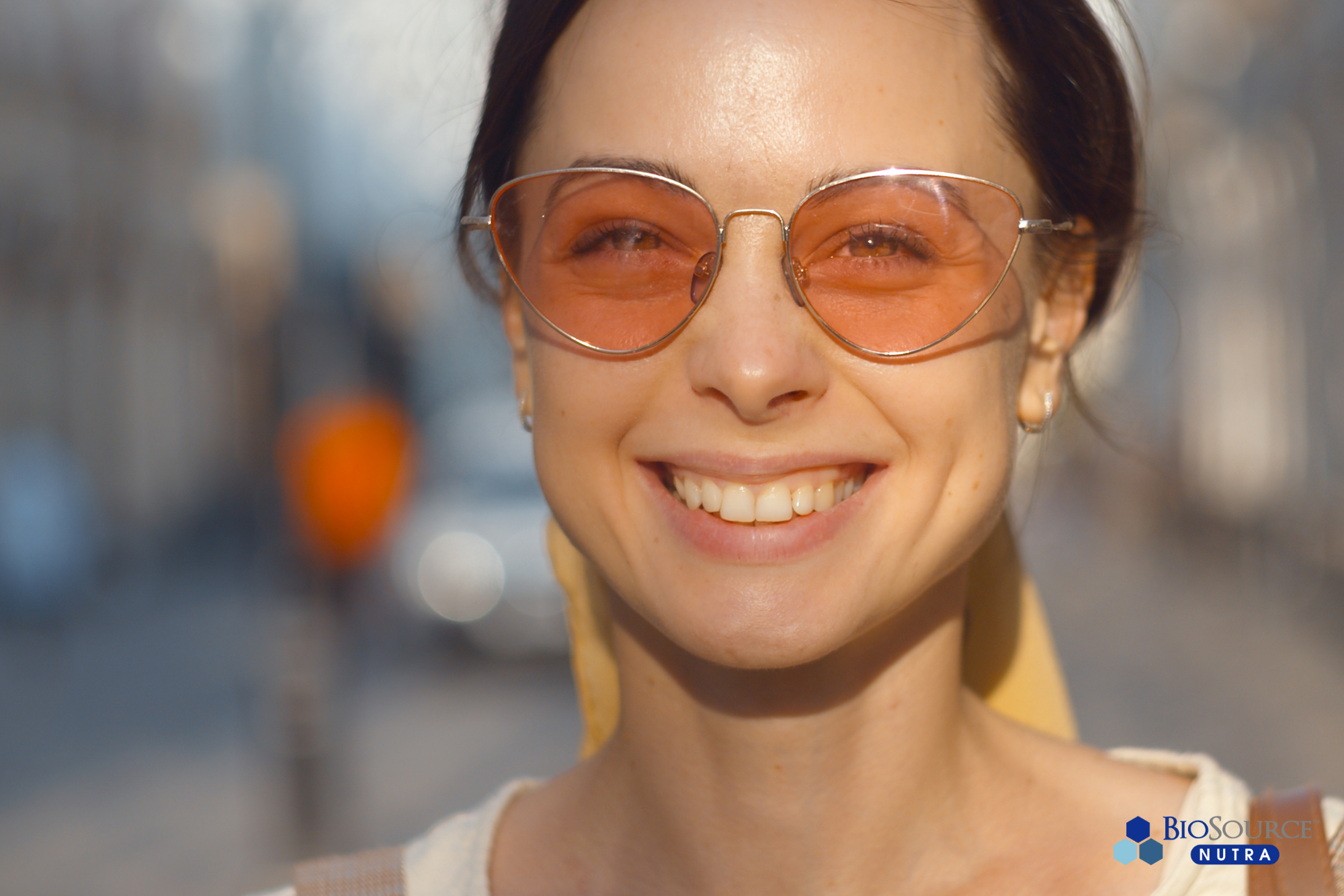 A young woman wearing orange tinted sunglasses is smiling in the sun