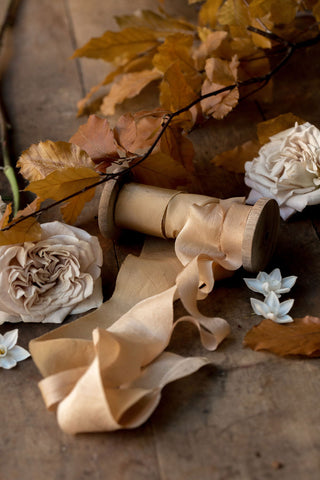 Gold and taupe silk ribbons on a spool with leaves and foliage on a table.