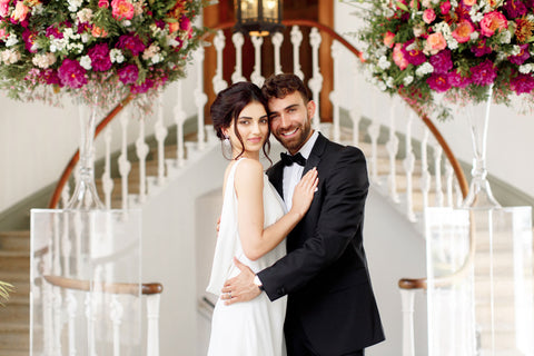 Bride and groom pose for photoshoot with bright floral display in background.