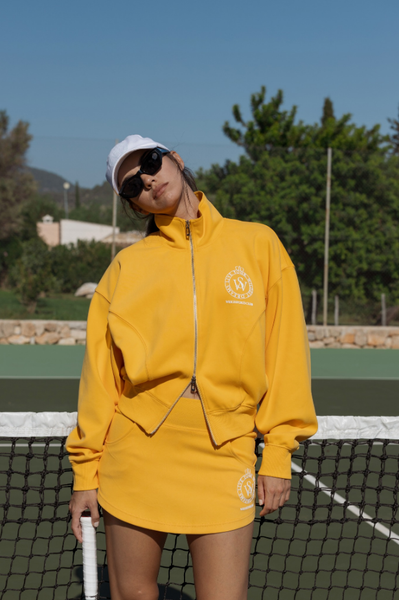 The model is wearing a loose yellow jacket, getting ready to play tennis outdoors.