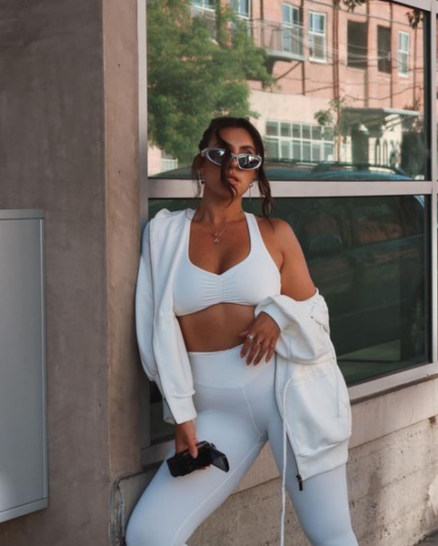 A woman is wearing an all-white athletic outfit paired with a cropped jacket and sunglasses, posing outdoors for a photo.