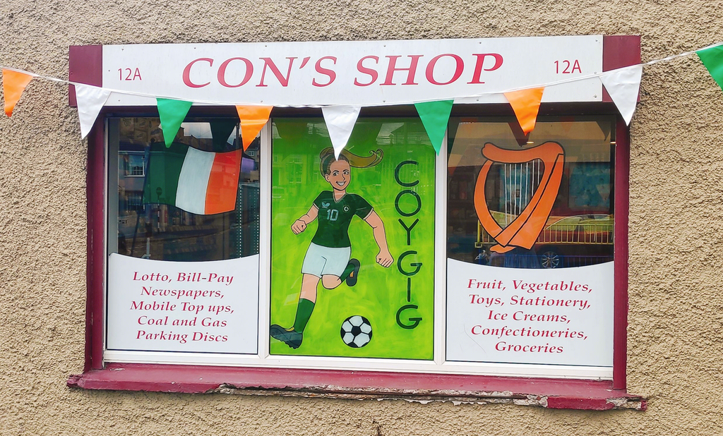 Photo of a window with a painting of the Irish tricolour, a harp and Denise O'Sullivan playing football. It also says COYGIG on the painting. The sign 'Con's Shop' is above the window. And Irish colour bunting frames the window at the top.