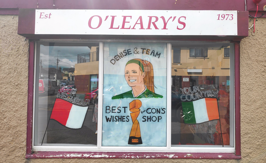 Photo of a second window painting of the Cork and Irish flags, and a portrait of Denise O'Sullivan with the Women's World Cup trophy under her. The text reads: Your Neighbour. Your Country. Denise & Team, Best Wishes from Con's Shop. Above the window is the sign 'O'Leary's, Est. 1973'.