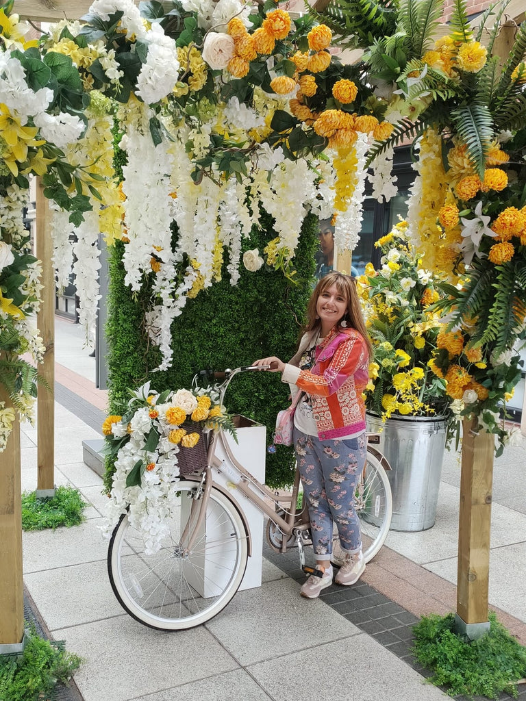 Photo of me posing with a white bike, surrounded by greenery and white, yellow and orange flowers. I'm wearing a pink and orange jacket with blue three quarter length pants with pink roses on them.