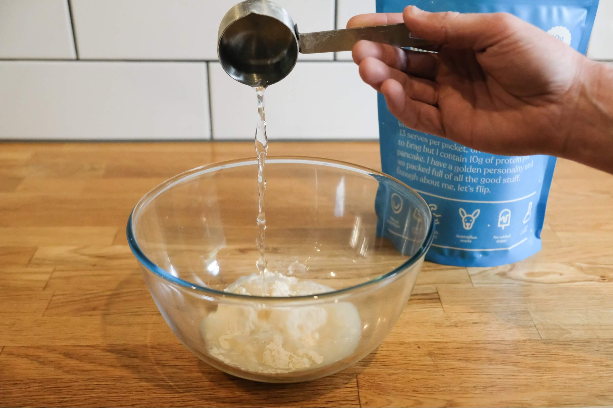 Person pouring pancake mix into a bowl from a measuring scoop, with mix package in background.