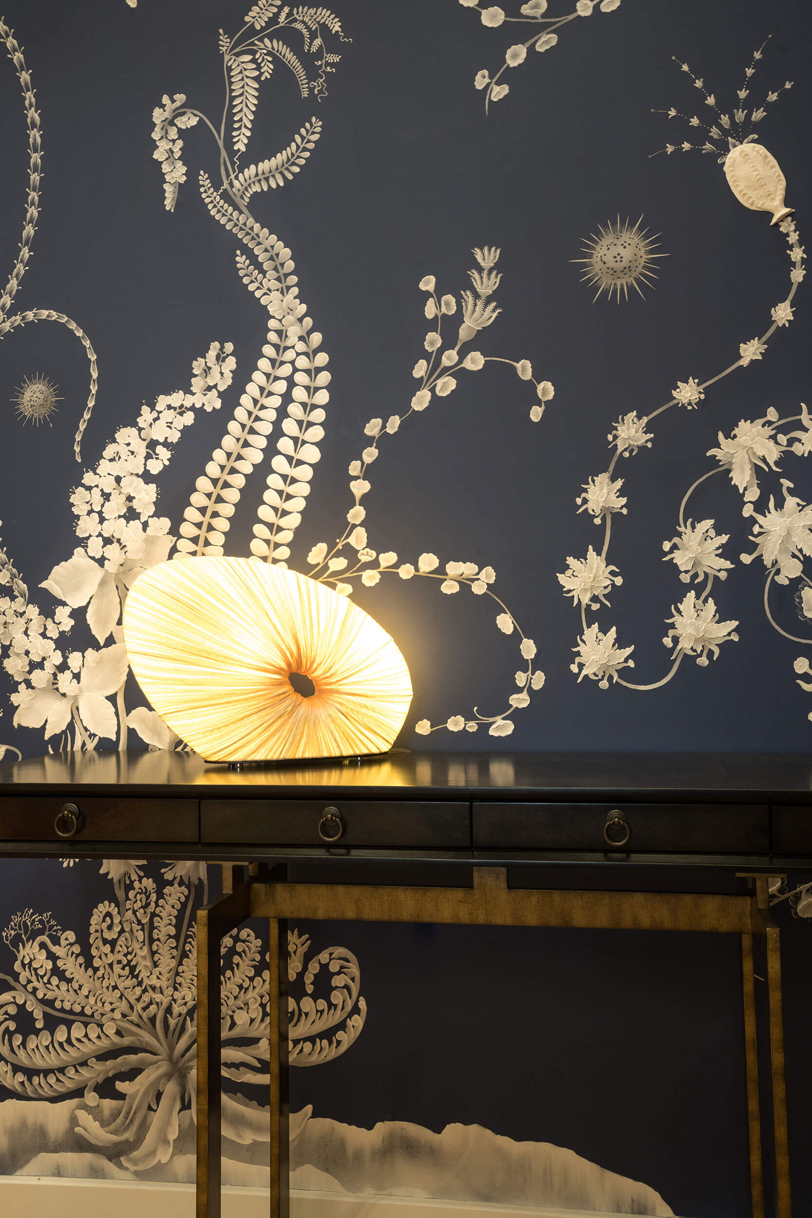 Doe Table Lamp in Cream silk on a dark wooden table against a dark blue wallpaper decorated with golden flower motifs