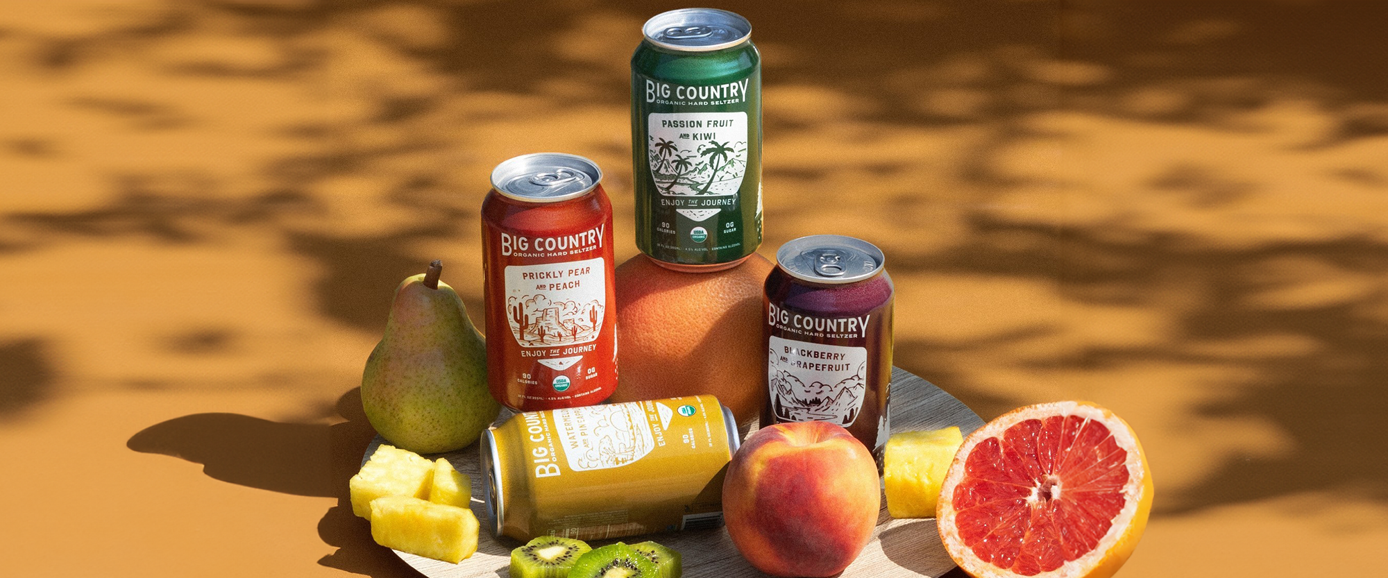 Hard seltzer cans among fruits on tan background with shadows.
