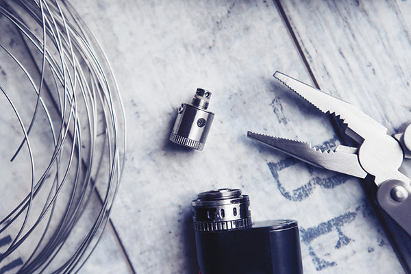 A vape, an atomizer, a coiled wire, and a pair of pliers lie on a wooden table. 