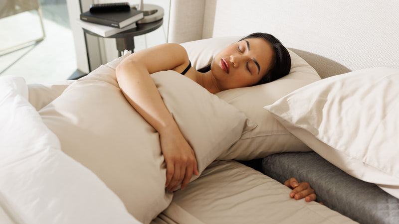 Photo of a female sleeper laying in the SONU mattress with her arm in the Comfort Channel while hugging a top pillow with cream sheets and pillowcases
