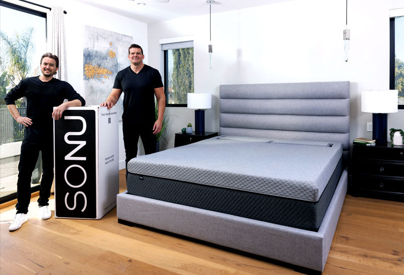 Photo of the SONU Sleep founders, Stason Strong & Brad Hall, next to their creation, the SONU Sleep System and its black and white box, in a bedroom with light grey wood floors.
