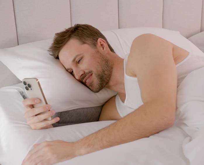 A male sleeper using his smart phone while being ergonomically supported in the SONU mattress with white sheets.