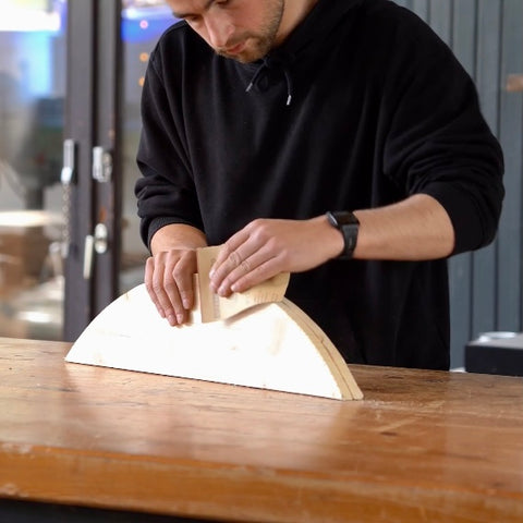 Eine Person steht in einer Werkstatt und schleift ein Halbrundes Holz. 