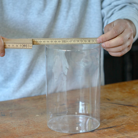Eine Person misst mit einem Zollstock den Durchmesser eines Glasgefäßes. 