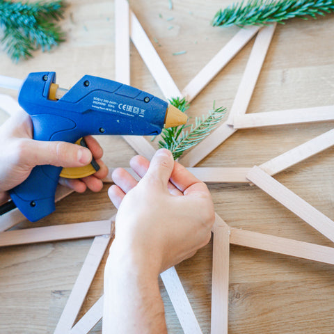 Ein Stern aus Holzleisten wird mit Tannengrün und einer Heißklebepistole beklebt. 