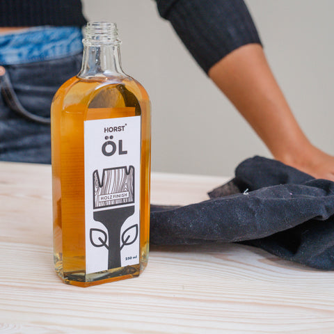 Eine vorgeschliffene Holzplatte soll mit einem Holzöl versiegelt werden. Auf einer Holzplatte steht eine Öl Flasche, liegt ein Baumwolltuch und ist eine Person sichtbar.