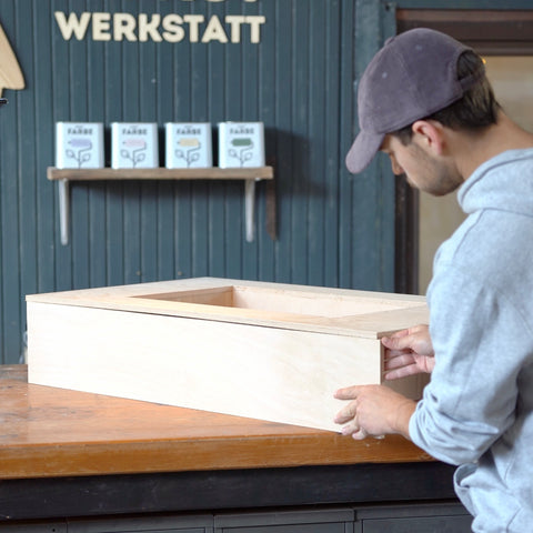 In einer Werkstatt wird ein Holzkasten zusammengebaut. Dabei schraubt eine Person mit einem Akkuschrauber zwei Holzplatten aneinander. 