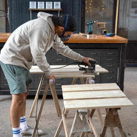 Eine Person schleift in einer Werkstatt auf mehreren Holzböcken einige Holzplatten.