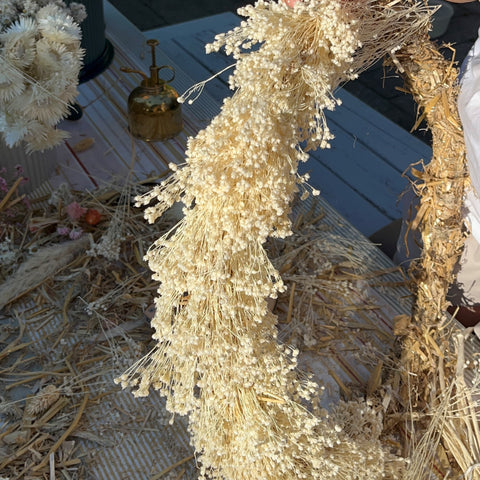 Ein Trockenblumenkranz ist bereits mit hellen Blumen umwickelt und wird weiter mit Hilfe von zwei Händen mit Blumen umwickelt. 