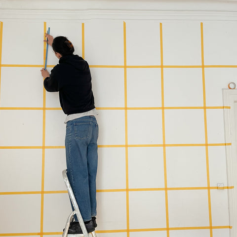 Eine Person steht auf einer Leiter und klebt mit Abklebeband ein Kariertes Muster auf die Wand. 