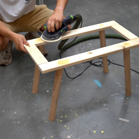 Eine Holzbank ohne Sitzfläche steht auf dem Boden. Eine Person kniet daneben und schleift mit einem Schleifgerät die Oberfläche an. 