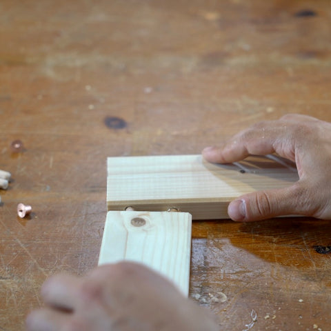 Auf einem Holztisch liegen zwei Holzleisten im rechten Winkel zueinander. Die Hände einer Person verbinden diese beiden Stücke.