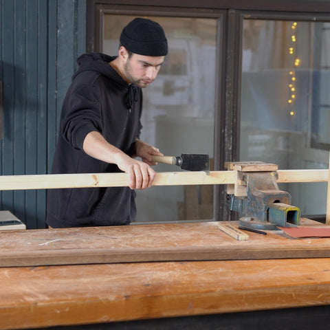 Eine Person steht in einer Werkstatt, hat eine Holzleiste eingespannt und sägt diese ab. 