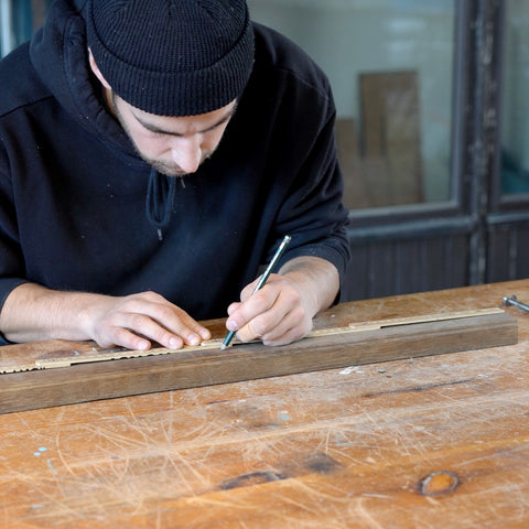 Eine Person arbeitet in seiner Werkstatt und zeichnet ein Maß auf eine Holzleiste. 