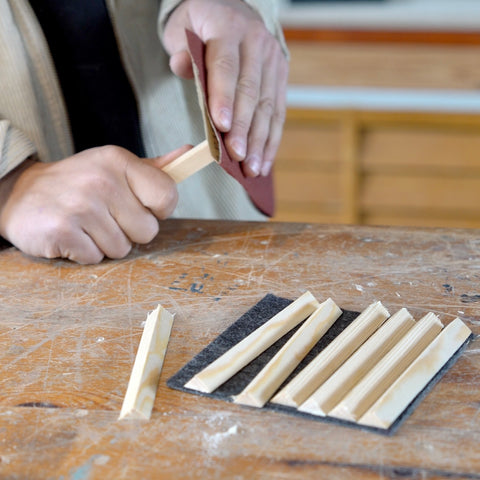 Auf einer Holzwerkbank werden Dreiecksleisten auf ein Stück Filz gelegt. Dabei schleift eine Person eine Holzleiste mit Schleifpapier ab. 