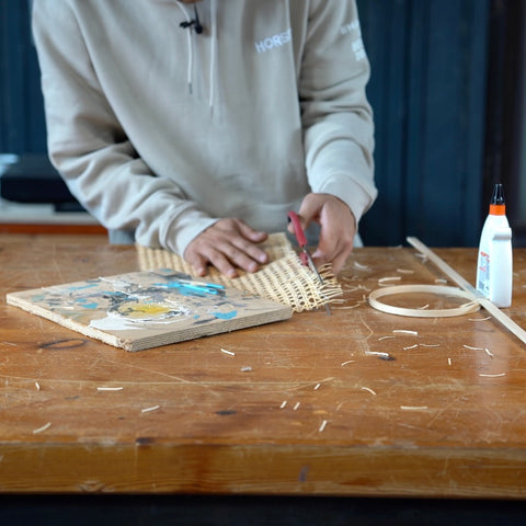 Auf einem Tisch schneidet eine Person ein Stück Wiener Geflecht mit einer Schere. Daneben liegt eine Holplatte, Holzleim und ein Stickrahmen.