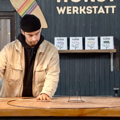 Eine Person misst mit Hilfe eines Zollstocks einen Abstand zwischen einem Metallkreis und einem Drahtgestell. Beides liegt auf einer Werkbank.