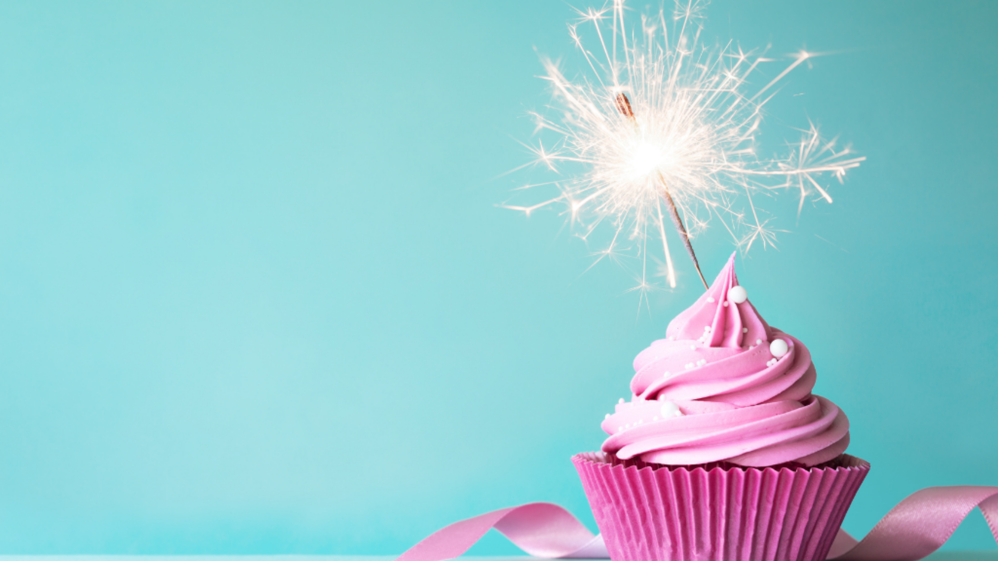 A pink cupcake with a lit sparkler on top sits on a turquoise background with a pink ribbon beside it.