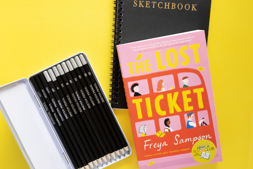 A book titled "The Lost Ticket" beside a sketchbook and pencils on a yellow surface.