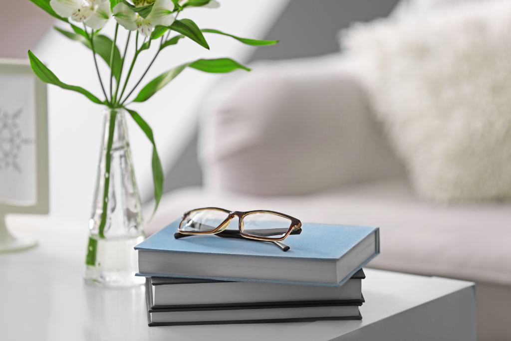 A stack of books with eyeglasses on top, beside a flower vase.