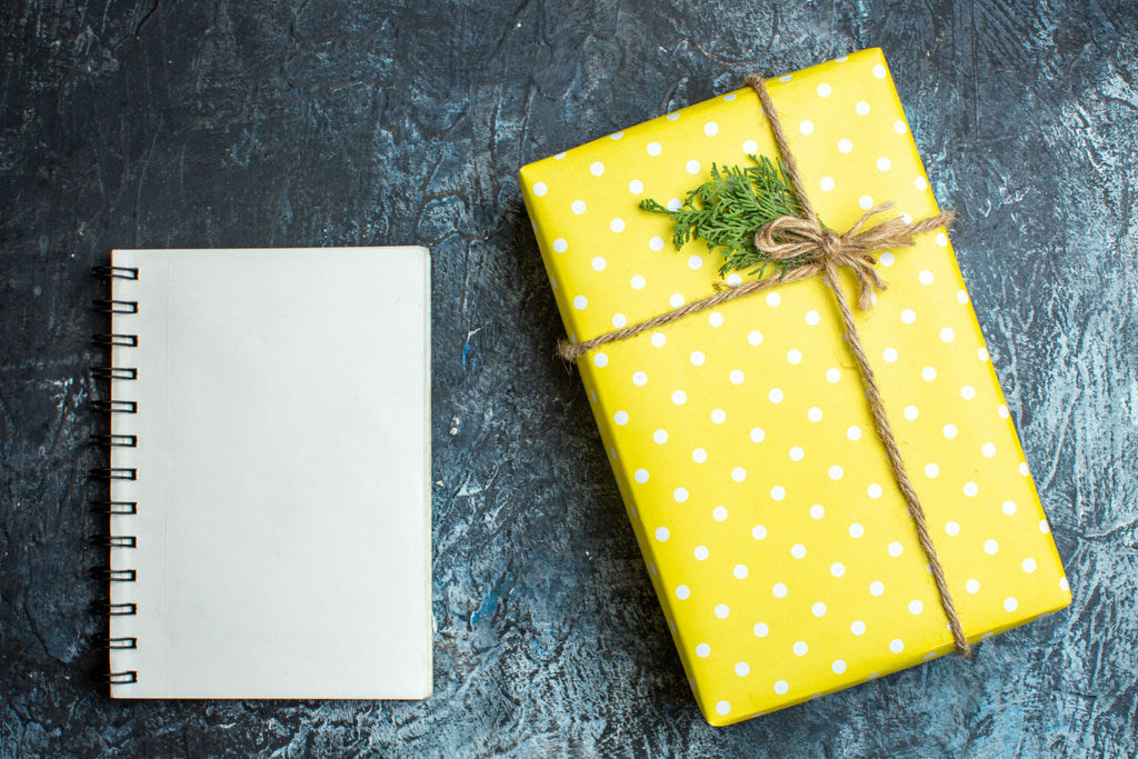 A cheerful yellow polka-dotted gift next to a blank notebook on a dark textured surface, tied with a rustic twine and a sprig of greenery, ready for a thoughtful note.