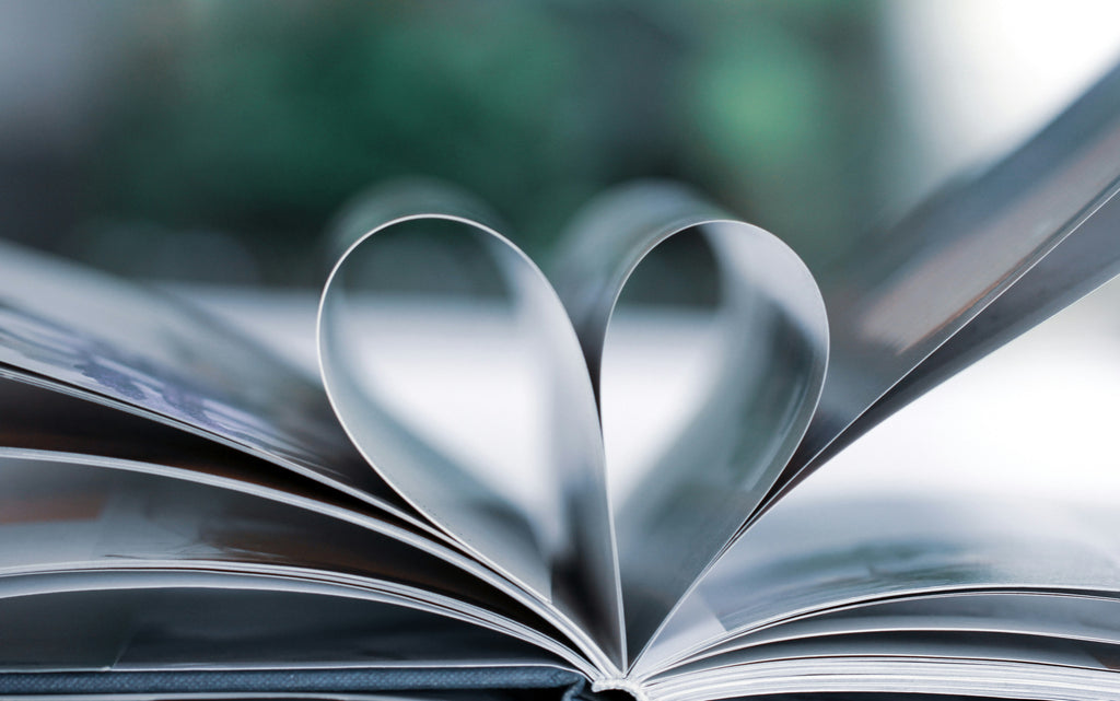 The pages of a book are turned down in the center to form a heart shape, symbolizing a love for reading. The focus on the heart-shaped pages creates a soft blur in the background, adding a gentle, romantic quality to the image.